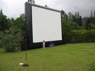 Une fillette se tient devant un grand écran gonflable blanc et noir installé sur une pelouse entourée de végétation luxuriante, avec des montagnes brumeuses en arrière-plan.