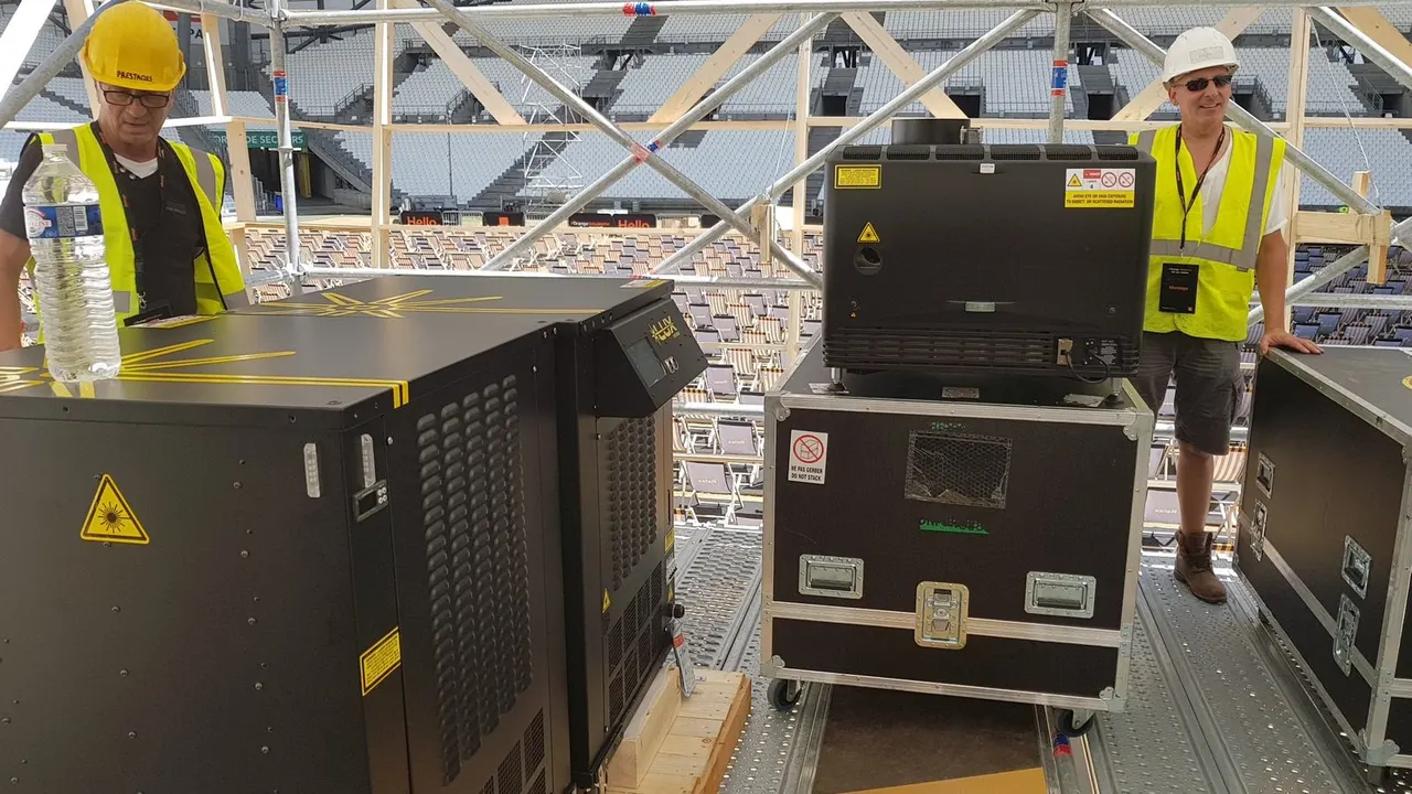 Deux techniciens en gilets jaunes et casques blancs installent du matériel de projection dans des flight cases sur une passerelle de stade vide
