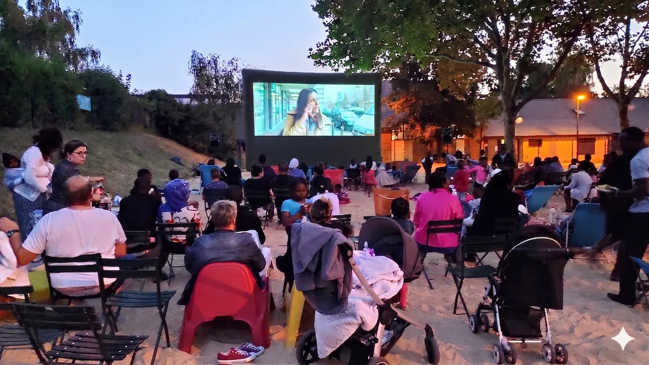 Un public nombreux assis sur des chaises et une poussette regarde un film projeté sur un écran gonflable au crépuscule dans un parc.