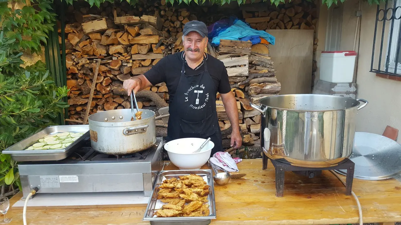 Un homme souriant portant un tablier et une casquette cuisine en extérieur devant un tas de bois, entouré de marmites et de plats de nourriture.