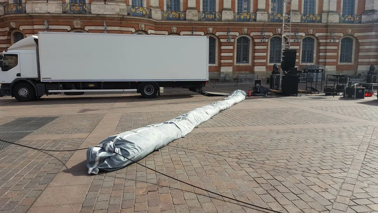Une longue housse grise contenant un écran gonflable plié est étendue sur les pavés d'une place devant un camion blanc et une façade en brique, en attente de montage.