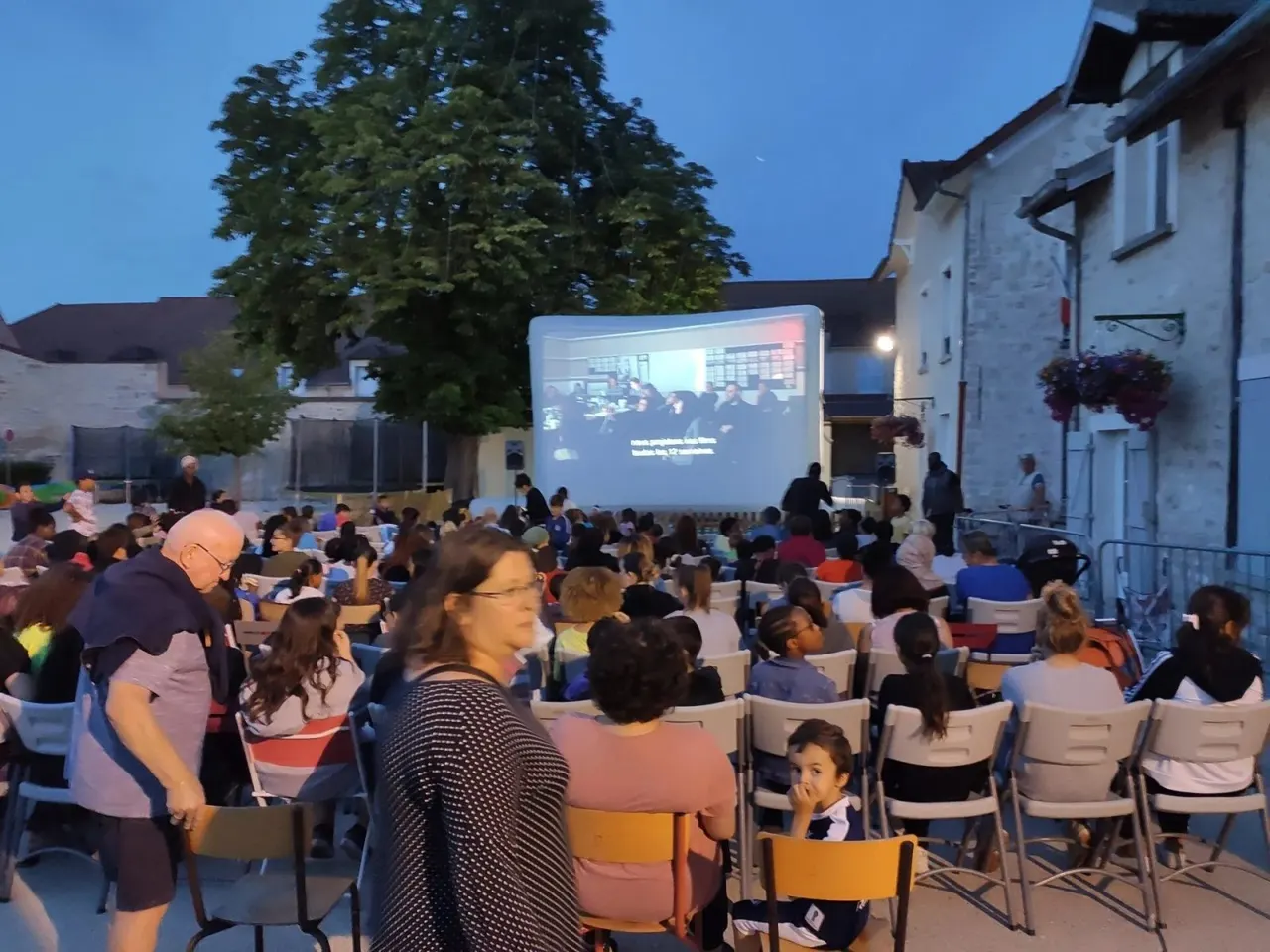 Une foule nombreuse assise sur des chaises regarde un film projeté sur un écran gonflable dressé sur une place de village, entourée de bâtiments en pierre à la tombée de la nuit.