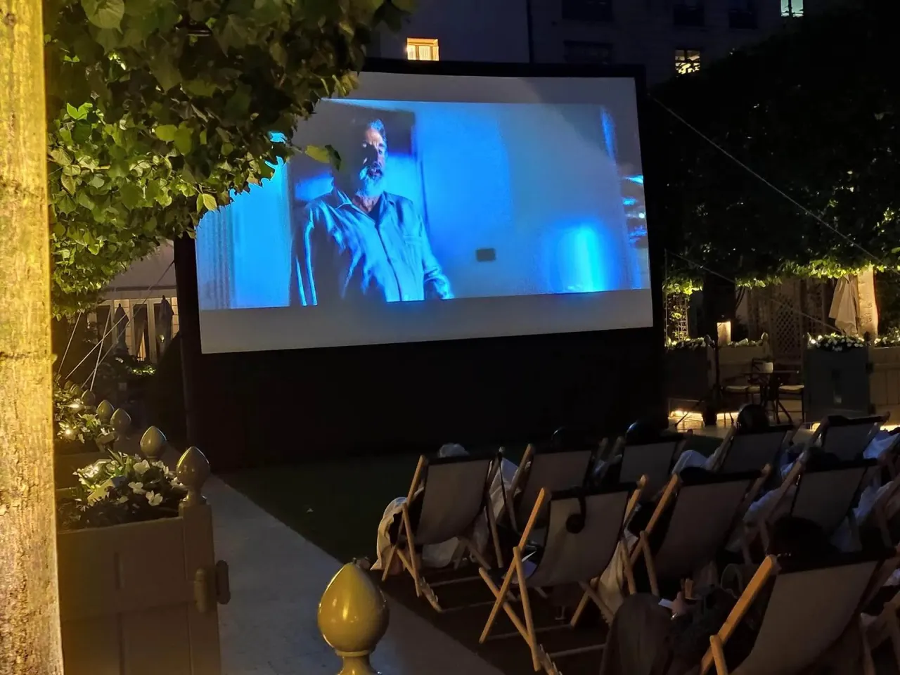 Un écran gonflable diffuse un film de nuit dans un jardin, entouré d'arbres, avec des chaises longues occupées par des spectateurs au premier plan.