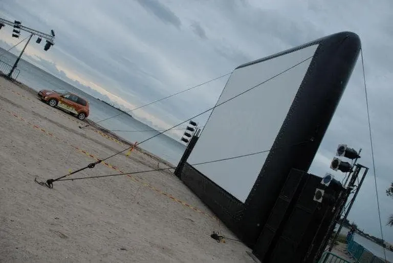 Un grand écran gonflable noir est haubané sur le sable en bord de mer, avec une voiture garée à proximité et des enceintes installées à droite.