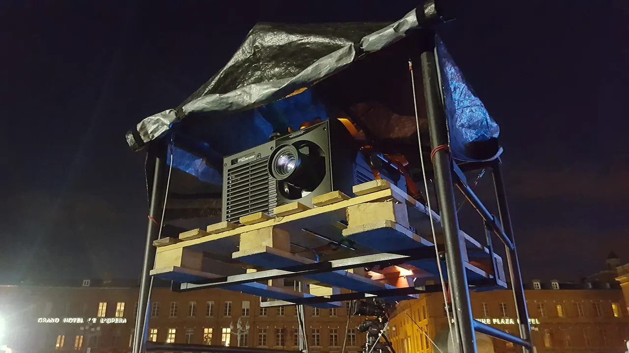 Gros plan sur un vidéoprojecteur professionnel posé sur des palettes en bois, protégé par une bâche, de nuit devant des façades d'hôtels éclairées.